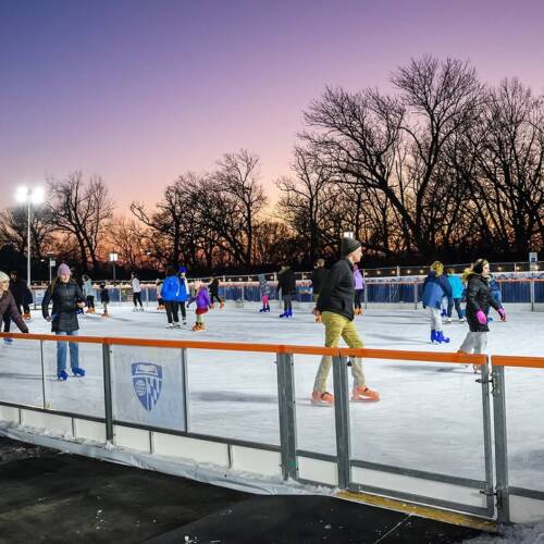 Skaters on an outdoor ice rink with a purple and pink sunset backdrop